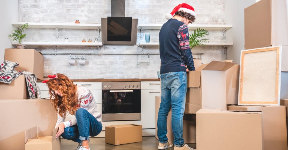 Um casal com gorros natalinos em meio a caixas, fazendo uma mudança no fim de ano.